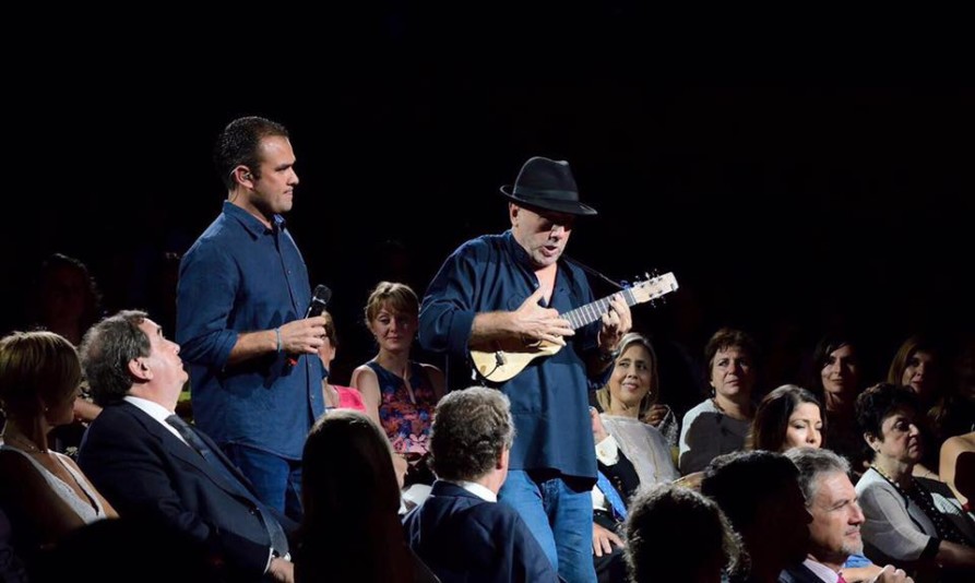 Un músico interpreta una pieza con un timple mientras camina entre el público durante un evento. A su lado, otro intérprete sostiene un micrófono. El público, atento, observa la actuación desde sus asientos.