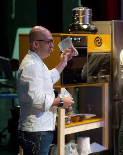 Un chef con chaqueta blanca sostiene una bolsa de vacío mientras ofrece una demostración culinaria en un escenario. Detrás de él, se observa un horno y utensilios de cocina profesionales.