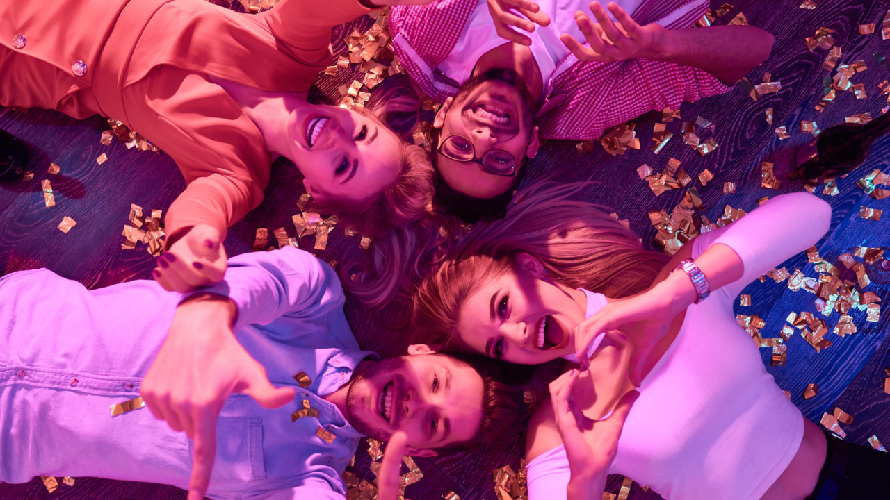 Top view at group of  happy young people gesturing hearts and thumbs up lying on floor among confetti and looking at camera during awesome house party.